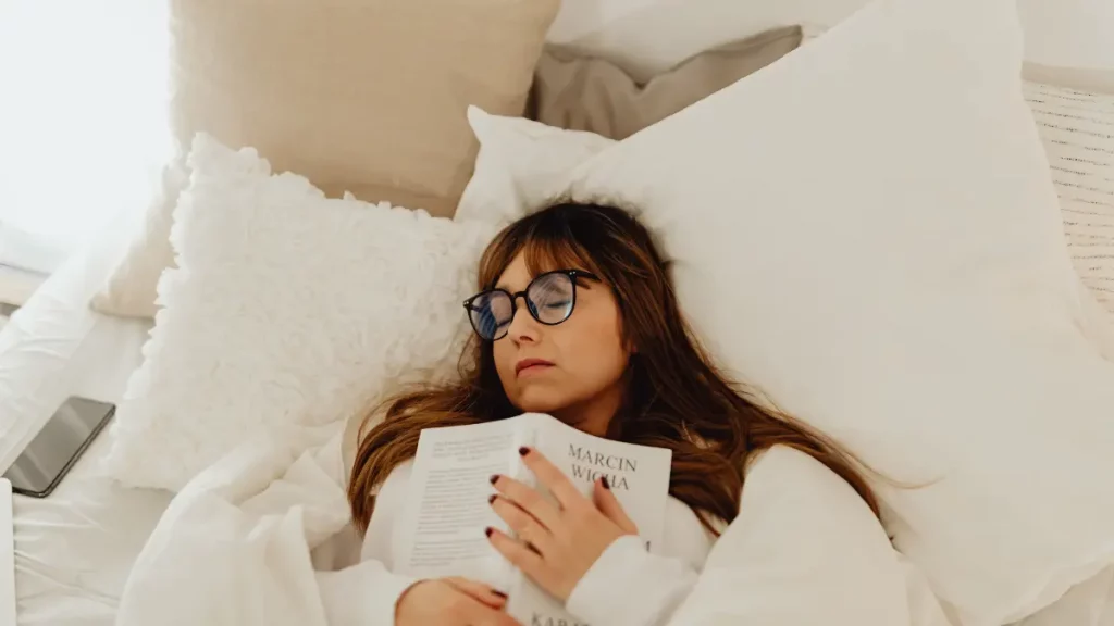 Frau liegt mit Brille und aufgeschlagenem Buch im hellen Bett und ist eingeschlafen, Sinnbild für Erschöpfung und Schlafstörungen in den Wechseljahren.