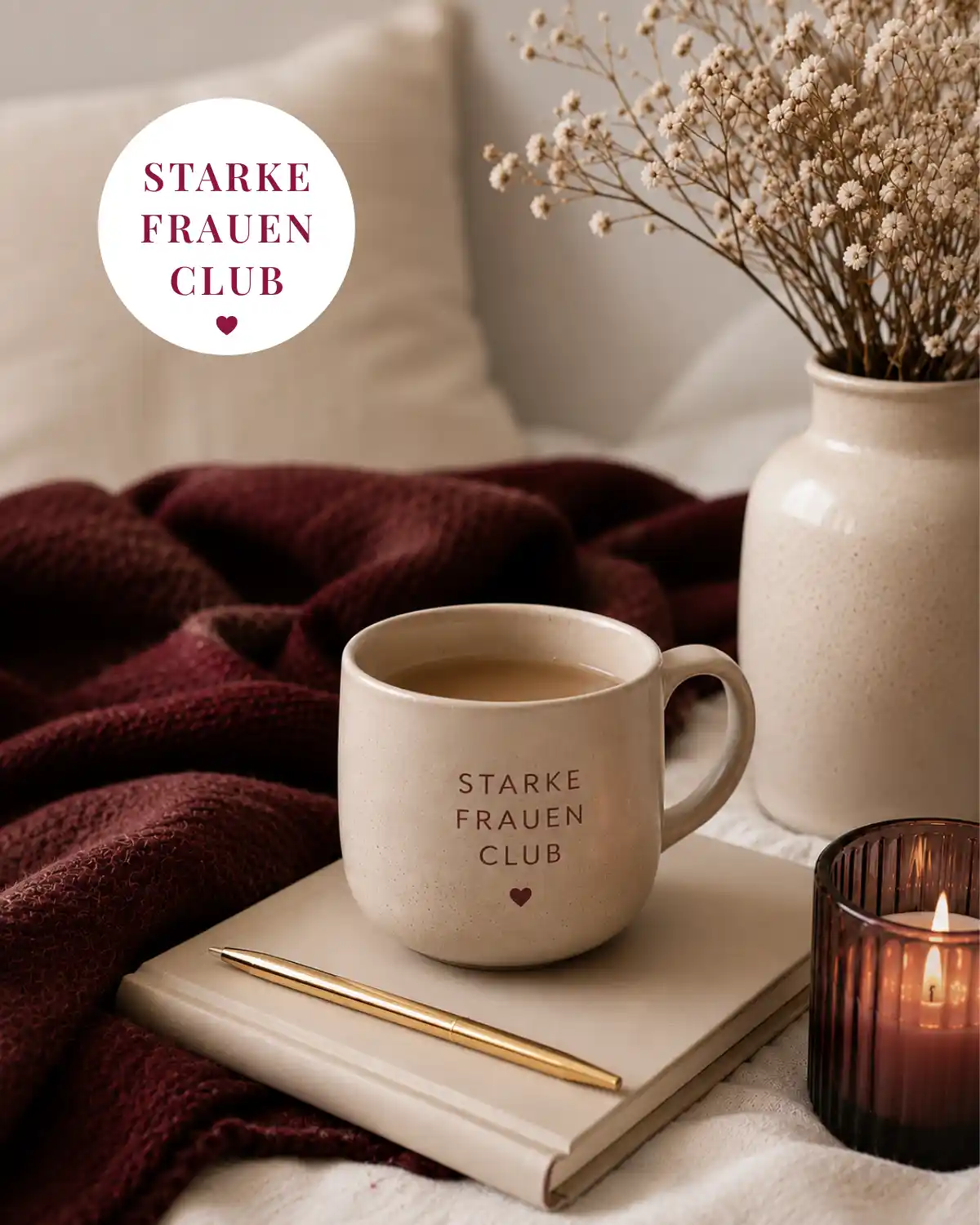 Beige Tasse mit der Aufschrift „Starke Frauen Club“ auf einem Notizbuch, daneben goldener Stift, Kerze und Trockenblumen